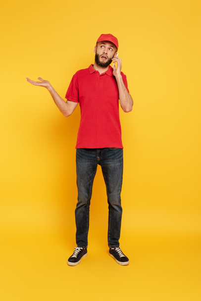 confused bearded delivery man in red uniform talking on smartphone on yellow - Photo, image