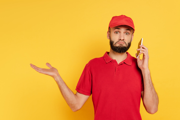 confused bearded delivery man in red uniform talking on smartphone on yellow - Photo, Image