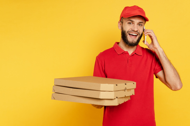 repartidor barbudo sonriente en uniforme rojo con cajas de pizza hablando en smartphone en amarillo
 - Foto, Imagen