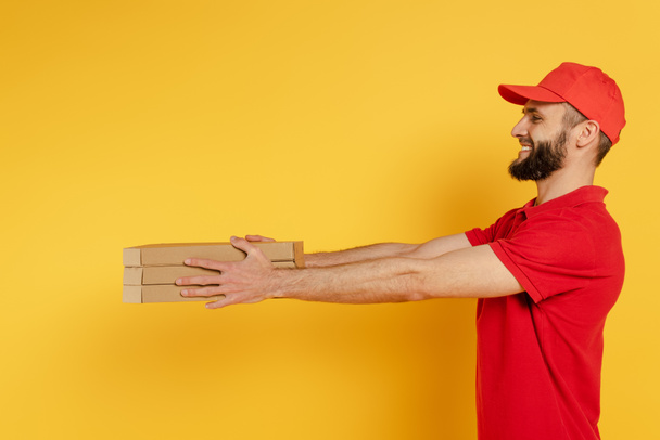side view of smiling bearded delivery man in red uniform giving pizza boxes on yellow - 写真・画像