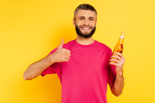happy bearded guy in pink t-shirt showing thumb up with beer on yellow - 写真・画像