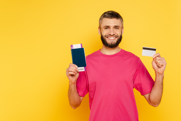 happy bearded guy in pink t-shirt with credit card and passport on yellow - Fotó, kép