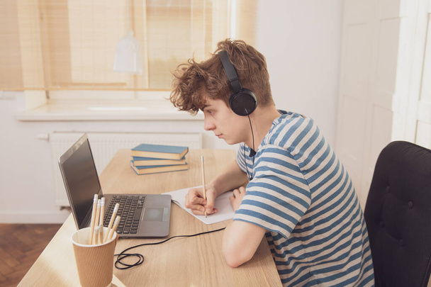 Egy kedves tinédzser fiú fülhallgatóban használja a laptopot, és a szobájában tanul az asztalnál. Egy ceruzával jegyzetel. Távolságtanulás a járványos koronavírus miatt - Fotó, kép