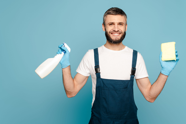 happy cleaner in uniform and rubber gloves holding detergent and sponge on blue background - Foto, afbeelding