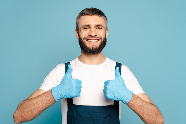happy cleaner in uniform en rubber handschoenen met duimen omhoog op blauwe achtergrond - Foto, afbeelding