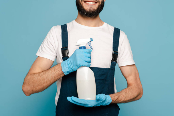 cropped view of happy cleaner in uniform and rubber gloves holding spray detergent on blue background - Foto, immagini
