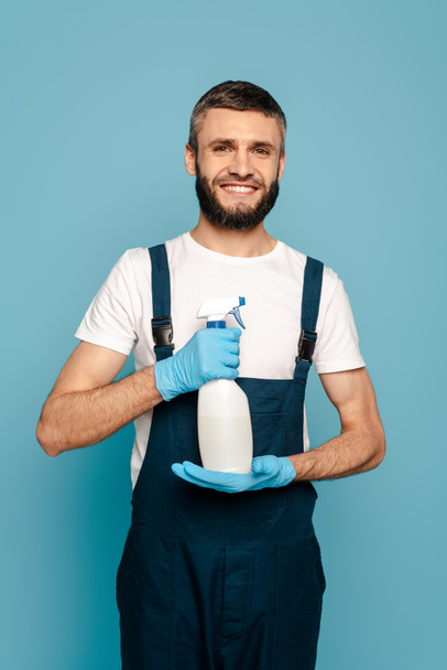 happy cleaner in uniform and rubber gloves holding spray detergent on blue background - Photo, Image