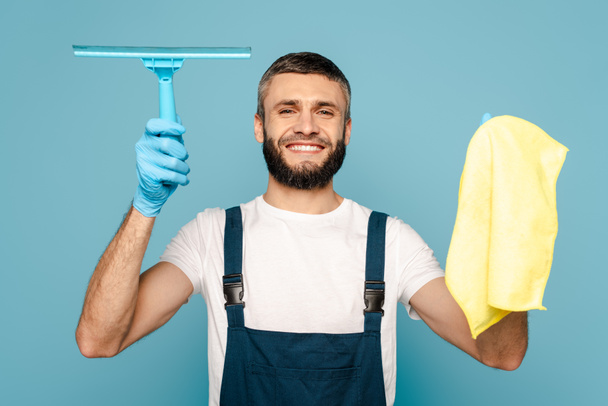 Üniformalı mutlu bir temizlikçi ve mavi arka planda bez ve cam sileceği tutan lastik eldivenler. - Fotoğraf, Görsel
