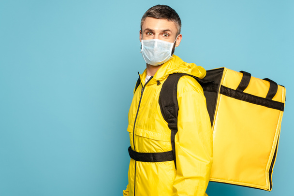 fattorino in maschera medica e uniforme gialla con zaino su sfondo blu
 - Foto, immagini