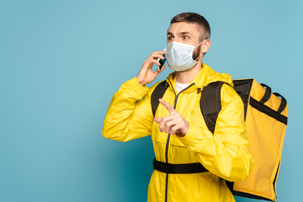deliveryman in medical mask and yellow uniform with backpack talking in smartphone and pointing with finger on blue background - Valokuva, kuva