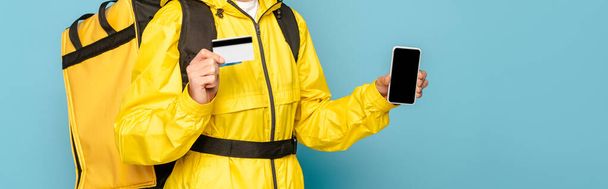 обрізаний вид доставляльника в жовтій формі з рюкзаком, що показує смартфон з порожнім екраном, тримаючи кредитну картку на синьому фоні
 - Фото, зображення