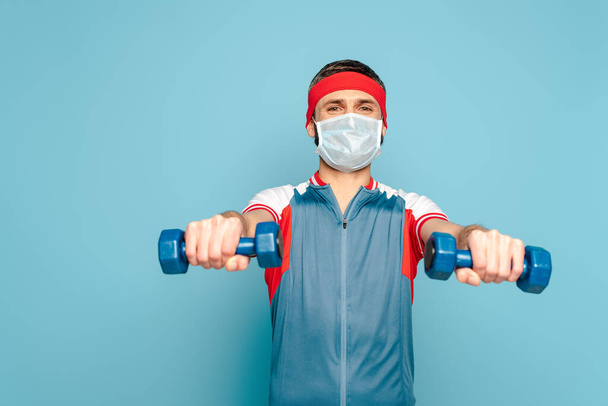 青い背景にダンベルで運動する医療用マスクのスタイリッシュなスポーツマン - 写真・画像
