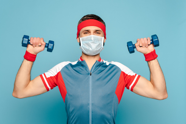 stylischer Sportler in medizinischer Maske beim Training mit Kurzhanteln auf blauem Hintergrund - Foto, Bild
