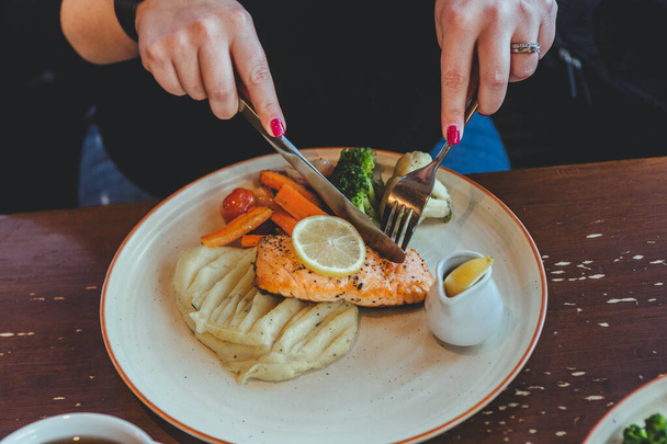 Руки женщины сидят за столом в кафе, едят стейк из красной рыбы и картофельное пюре с овощами на белой тарелке
 - Фото, изображение