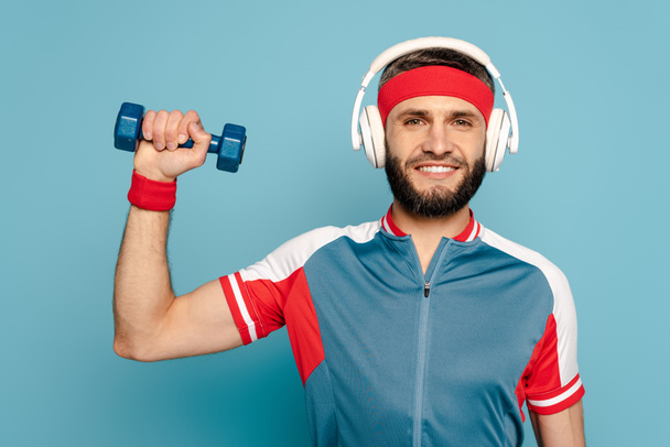 happy stylish sportsman in headphones exercising with dumbbell on blue background - 写真・画像