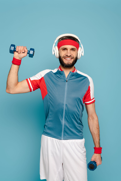 happy stylish sportsman in headphones exercising with dumbbells on blue background - 写真・画像