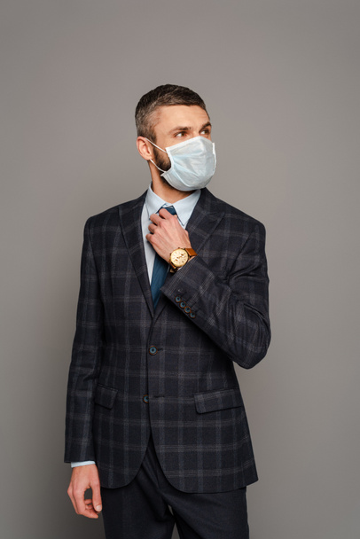 όμορφος επιχειρηματίας με ιατρική μάσκα που προσαρμόζει τη γραβάτα σε γκρι φόντο - Φωτογραφία, εικόνα