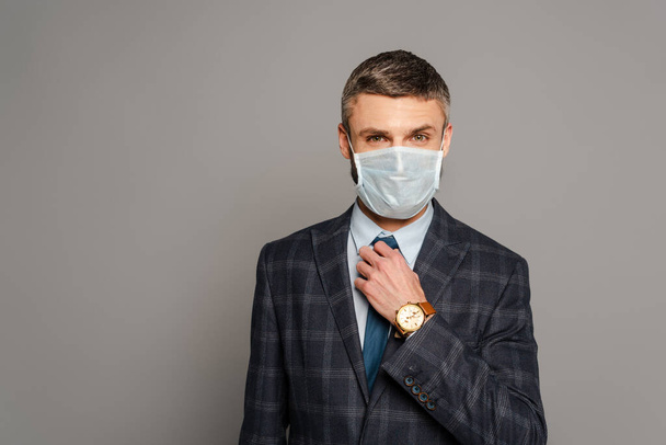 όμορφος επιχειρηματίας με ιατρική μάσκα που προσαρμόζει τη γραβάτα σε γκρι φόντο - Φωτογραφία, εικόνα