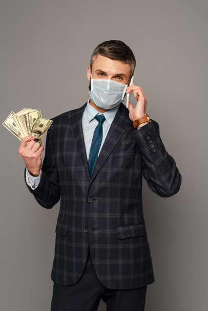 όμορφος επιχειρηματίας με ιατρική μάσκα με χρήματα μιλώντας σε smartphone σε γκρι φόντο - Φωτογραφία, εικόνα