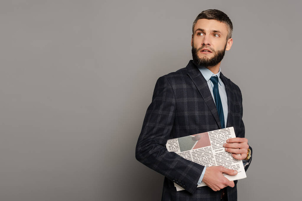 smiling handsome bearded businessman in suit with newspaper on grey background - Fotoğraf, Görsel