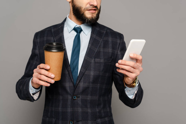 cropped view of bearded businessman in suit with coffee to go using smartphone on grey background - 写真・画像