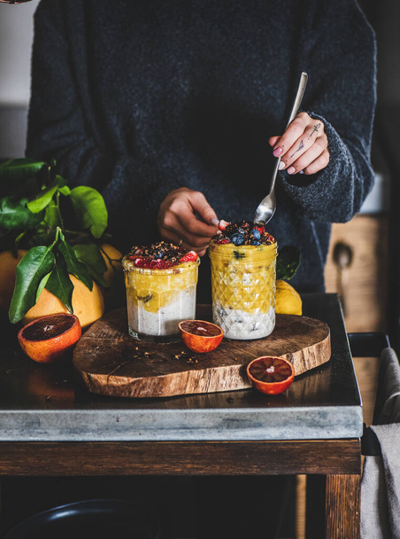 Woman eating overnight oat bircher muesli with mango smoothie, buckwheat granola and fresh berries from glass with spoon over grey concrete kitchen counter. Healthy, clean eating concept - Zdjęcie, obraz