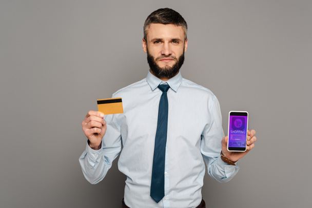 Yakışıklı sakallı iş adamı, elinde kredi kartı ve gri arka planda online alışveriş uygulaması olan akıllı telefon tutuyor. - Fotoğraf, Görsel