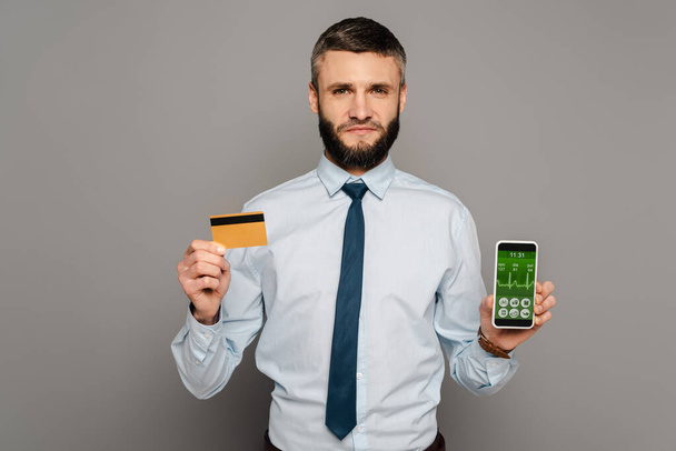 guapo barbudo hombre de negocios con tarjeta de crédito y teléfono inteligente con aplicación de salud sobre fondo gris
 - Foto, Imagen