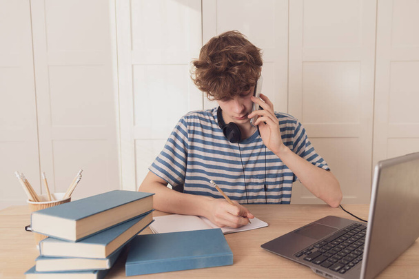 Teenager talking on a cell phone, learning using a laptop.A thoughtful kid at his desk is thinking about learning.Distance learning because of the epidemic coronavirus - Photo, Image