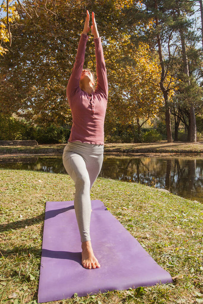 Młoda kobieta ćwiczy jogę na macie w parku nad jeziorem w piękny jesienny dzień rano. - Zdjęcie, obraz
