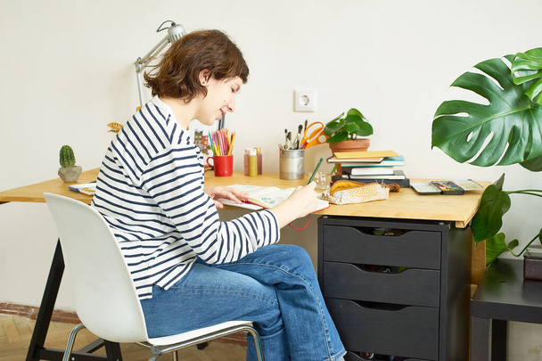 Female artist at her workplace working from home. Woman dressed in jeans and striped shirt sitting at the table and drawing illustration.  - Fotografie, Obrázek