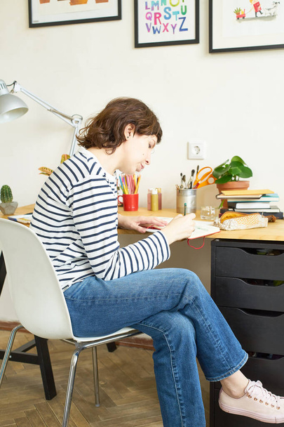 Female artist at her workplace working from home. Woman dressed in jeans and striped shirt sitting at the table and drawing illustration.  - Фото, зображення
