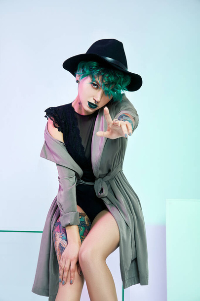 Woman in hat with creative green coloring hair and makeup, toxic strands of hair. Bright color curly hair on the girl head, professional makeup. Woman with tattoo in cloak - 写真・画像