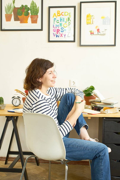 Female artist at her workplace working from home. Young woman dressed in jeans and striped shirt sitting at the table, smiling and creating an illustration.  - Foto, afbeelding