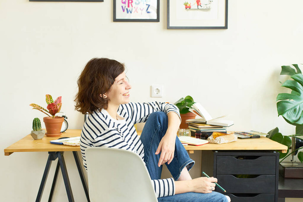 Female artist at her workplace working from home. Woman dressed in jeans and striped shirt sitting at the table and drawing illustration. Looking for inspiration concept. - Photo, image