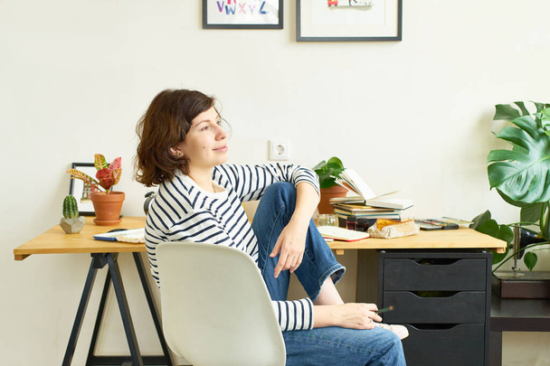 Female artist at her workplace working from home. Woman dressed in jeans and striped shirt sitting at the table and drawing illustration.  - Foto, imagen