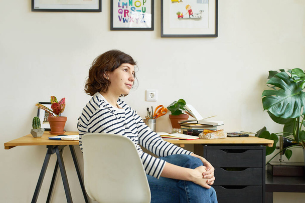 Female artist at her workplace working from home. Woman dressed in jeans and striped shirt sitting at the table and drawing illustration. Looking for inspiration concept. - Foto, immagini