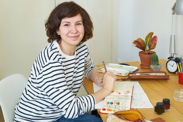 Artista femenina en su lugar de trabajo trabajando desde casa. Mujer joven vestida con jeans y camisa a rayas sentada a la mesa, sonriendo y creando una ilustración
.  - Foto, Imagen