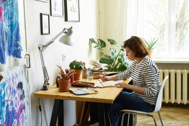 Female artist at her workplace working from home. Woman dressed in jeans and striped shirt sitting at the table and drawing illustration.  - Photo, Image