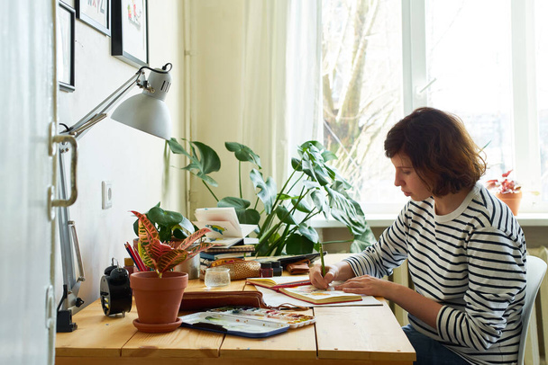 Artista femenina en su lugar de trabajo trabajando desde casa. Mujer vestida con jeans y camisa a rayas sentada a la mesa y dibujando ilustraciones
.  - Foto, imagen
