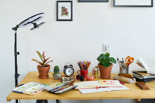 Artist's workplace for working from home with watercolor paints, brushes and sketchbooks. Place for design, illustration and creativity. Blogger ring light standing next to the table. - Photo, Image