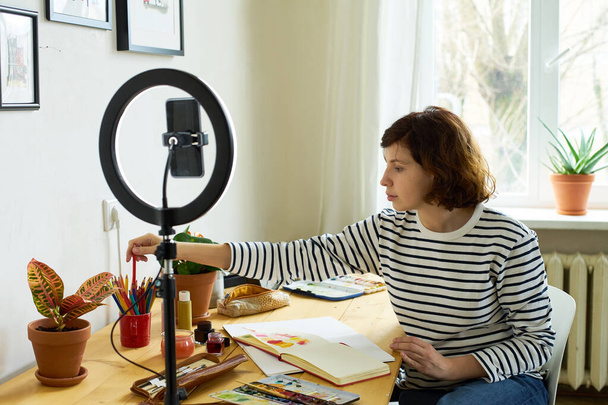 Female artist filming design workshop at home for online education. Working from home making master-class using ring lighting. Cozy artist's studio.  - 写真・画像