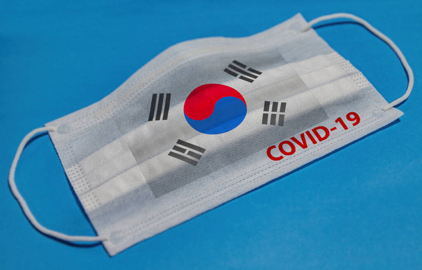 Medical face mask with  flag of South Korea and COVID-19 text on blue background. Concept of coronavirus quarantine. - Foto, imagen