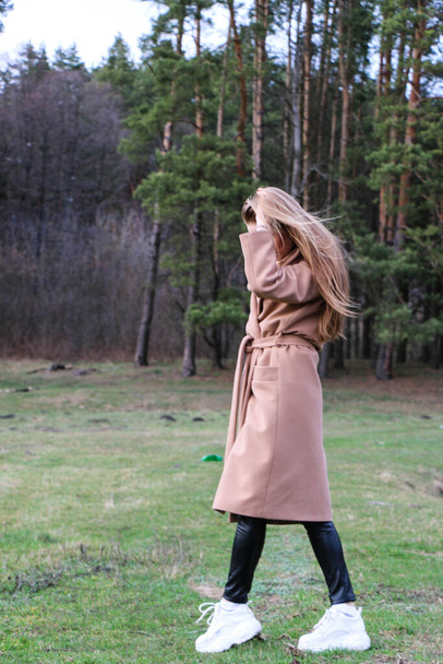beautiful girl, blonde with long hair, walks in the spring near the forest in a brown coat., serenity and freedom - Foto, Imagen