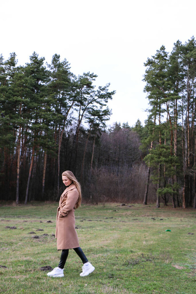 beautiful girl, blonde with long hair, walks in the spring near the forest in a brown coat., serenity and freedom - Fotó, kép