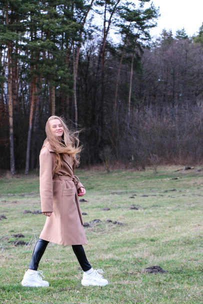 beautiful girl, blonde with long hair, walks in the spring near the forest in a brown coat., serenity and freedom - Foto, afbeelding