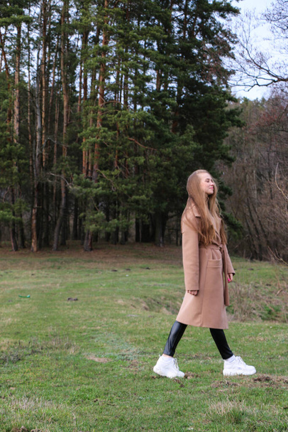 beautiful girl, blonde with long hair, walks in the spring near the forest in a brown coat., serenity and freedom - Zdjęcie, obraz