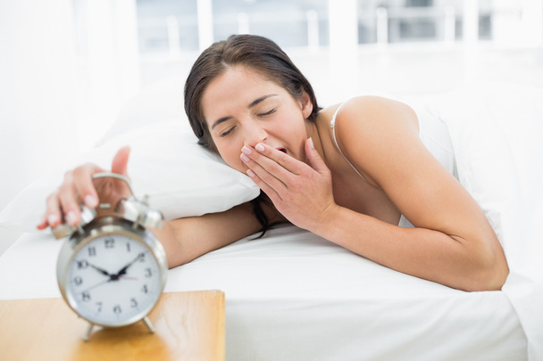 Woman yawning while extanding hand to alarm clock - Fotó, kép