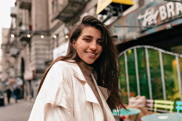 Femme européenne avec sourire heureux et cheveux foncés souriant à la caméra et marchant avec des émotions heureuses sur fond de centre-ville
. - Photo, image