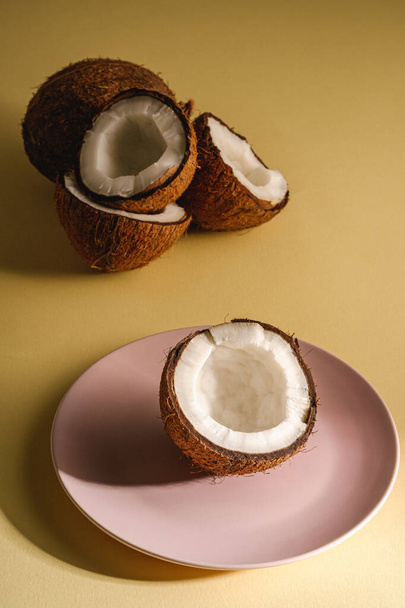 Coconut half in pink plate with nut fruits on cream yellow plain background, abstract food tropical concept, angle view - Φωτογραφία, εικόνα